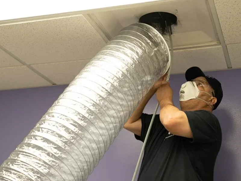 Technician performing air duct cleaning, wearing a mask, removing debris from ductwork to improve indoor air quality and support HVAC efficiency.