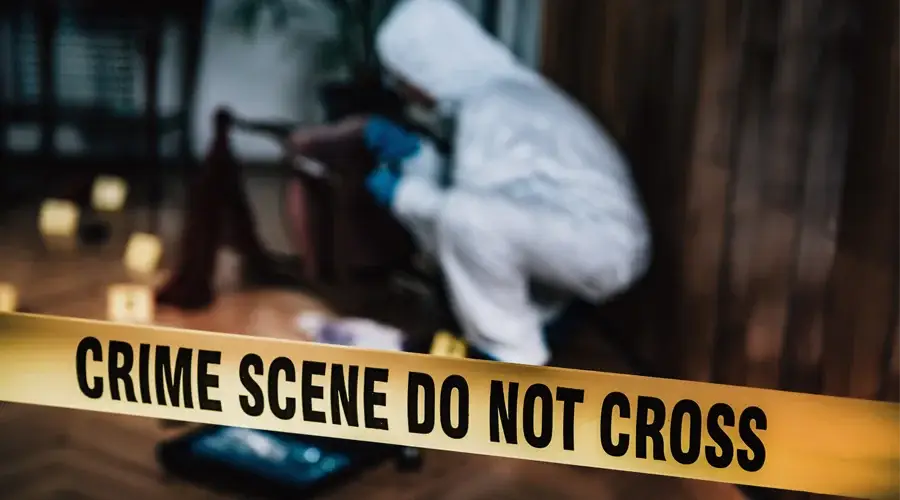 Person in protective gear investigating a crime scene, with yellow "CRIME SCENE DO NOT CROSS" tape in the foreground, emphasizing the need for professional trauma cleanup and safety measures in commercial properties.
